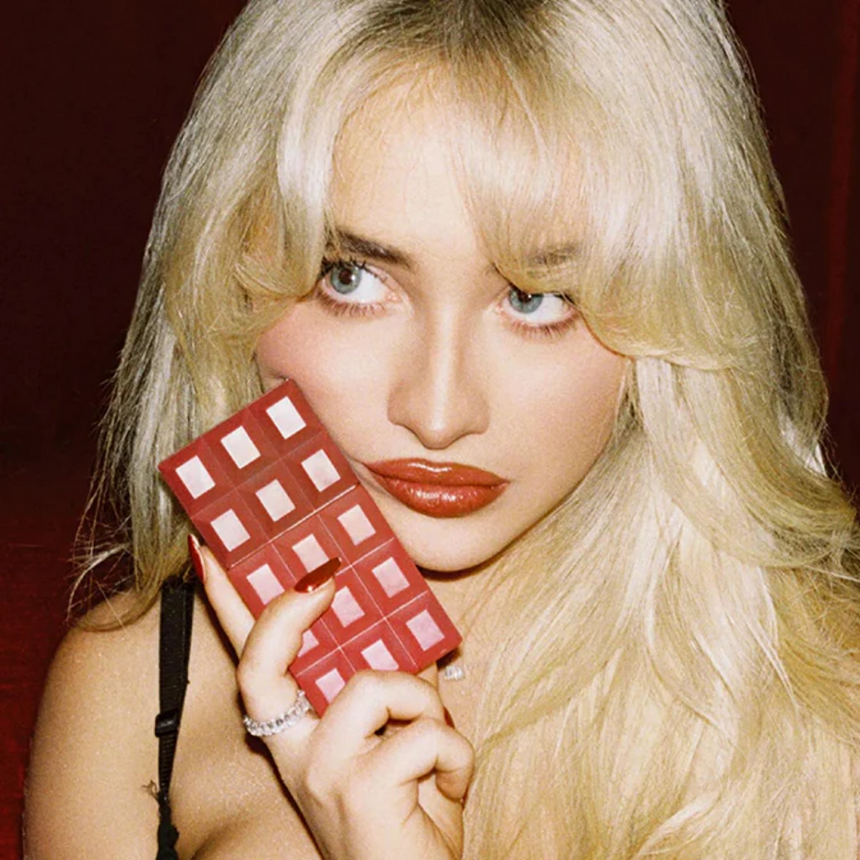 A spotlighted woman holding the Sabrina Carpenter Cherry Baby Fragrance in a dark red setting.