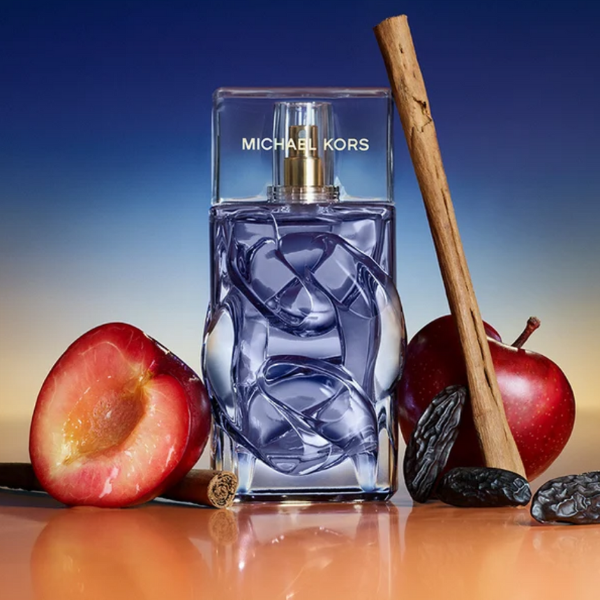 Michael Kors Pour Homme fragrance bottle surrounded by plum, apple, cinnamon and tonka beans against a blue background.