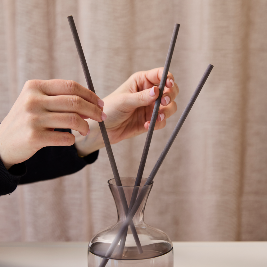 Hands arranging dark diffuser reeds in a glass vessel against a soft neutral backdrop.