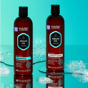 Hask Haircare Argan Oil Shampoo and Conditioner bottles displayed on a reflective turquoise surface with bubbles.