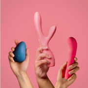 Hands holding three colourful sexual wellness toys against a pink studio backdrop.