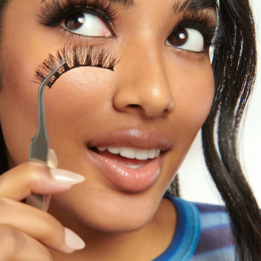 Close-up of a smiling woman holding a KISS lash strip and lash applicator against a white studio backdrop.