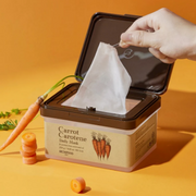 Hand pulling a SkinFood Carrot Carotene Calming Water Pad from an open tub, styled with fresh carrots against a warm orange background.