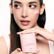 Woman holding House Of Hur Purifying Cleansing Balm with a product swatch across her cheek against a white backdrop.