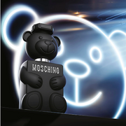 Black Moschino teddy bear fragrance bottle displayed in front of a glowing neon bear outline on a dark blue background.
