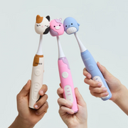 Three Ordo electric toothbrushes in beige, pink and blue with playful animal character brush heads, held against a light backdrop.
