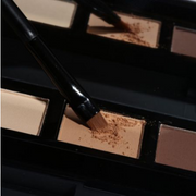 HD Brows Eyeshadow palette with a makeup brush applying a warm brown shade against a dark, glossy backdrop.