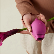 Hand holding Smile Makers The Poet Clitoral Suction Vibrator with a flower against a light beige backdrop.