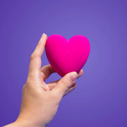 Hand holding So Divine Je T'Aime Couple's Heart Vibrator against a purple backdrop.