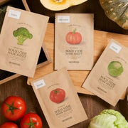 Skin Food Sous Vide sheet masks in broccoli, pumpkin, tomato and cabbage varieties, arranged with fresh vegetables on a wooden surface.