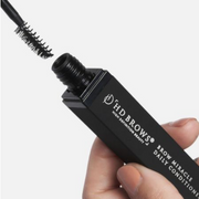 Hand holding HD Brows Brow Miracle Daily Conditioner in a white studio background.