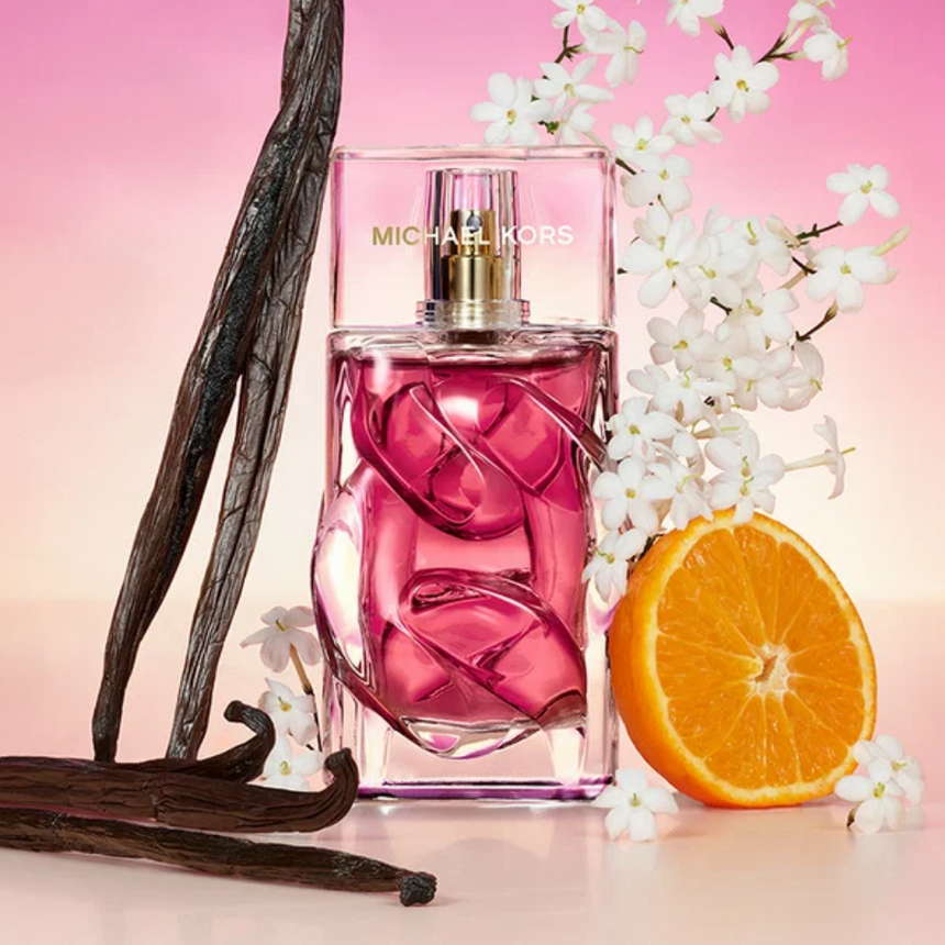 Michael Kors Pour Femme Pink fragrance bottle surrounded by vanilla pods, white flowers and orange against a pink background.
