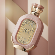 Pink and gold luxury fragrance bottle with an ornate gold label, displayed against a soft blush and champagne geometric background.