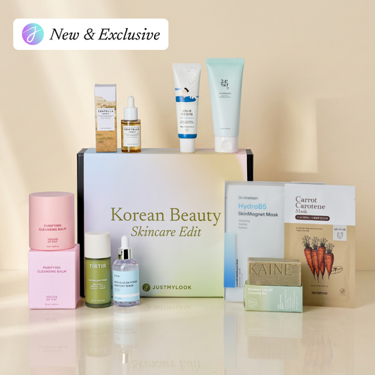 A curated Korean beauty skincare gift set arranged around a central white box labeled 'Korean Beauty Skincare Edit' by Justmylook. The set includes pink and green skincare jars, a small amber dropper bottle, a white tube, a pale blue tube, a sheet mask packet, and a carrot-face mask pack, all on a warm beige reflective surface with a 'New & Exclusive' badge in the top-left corner.